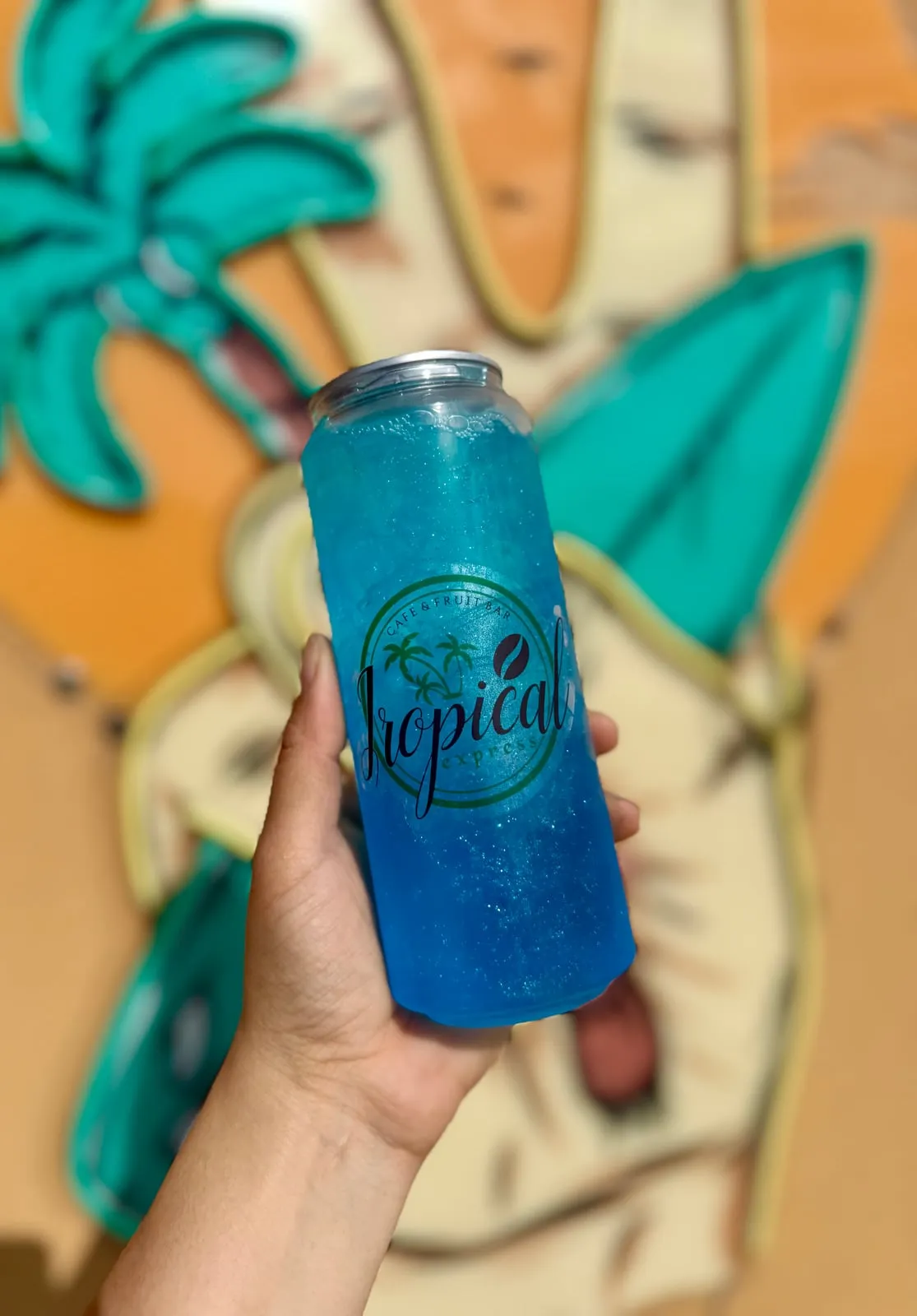 A sealed clear Tropical Express can of Ocean Mist — a vivid blue lotus and blue-razz refresher shimmering with edible glitter — held in front of the shop’s painted palm-tree mural