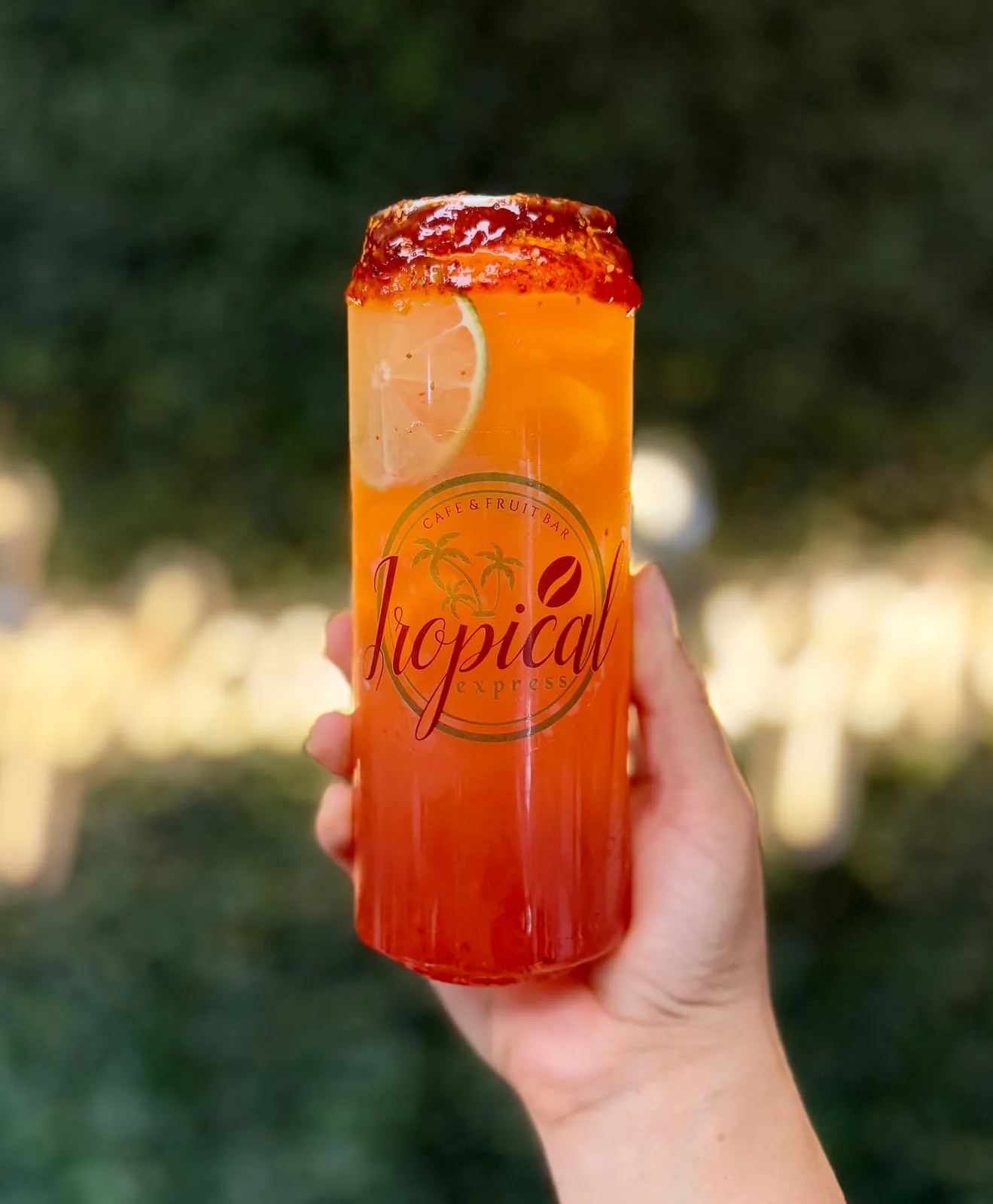 A second sealed clear Tropical Express can of a Cantarito — a sunset-orange grapefruit, watermelon and pineapple refresher with a chili-lime chamoy rim and a lime wheel — held in the sun