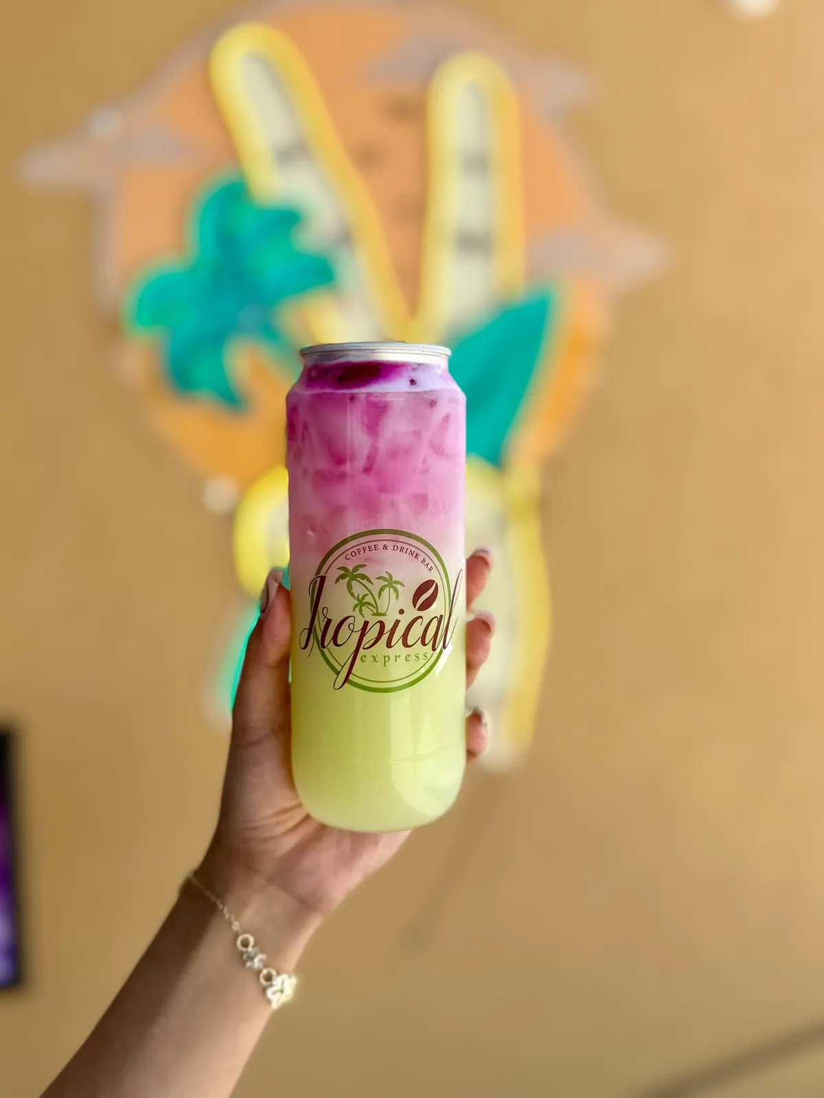 A sealed clear Tropical Express can of Dragon Paradise — a layered drink, magenta dragon fruit on top fading into a pale green cantaloupe and kiwi-passion base with creamy coconut milk — held in front of the painted palm sign