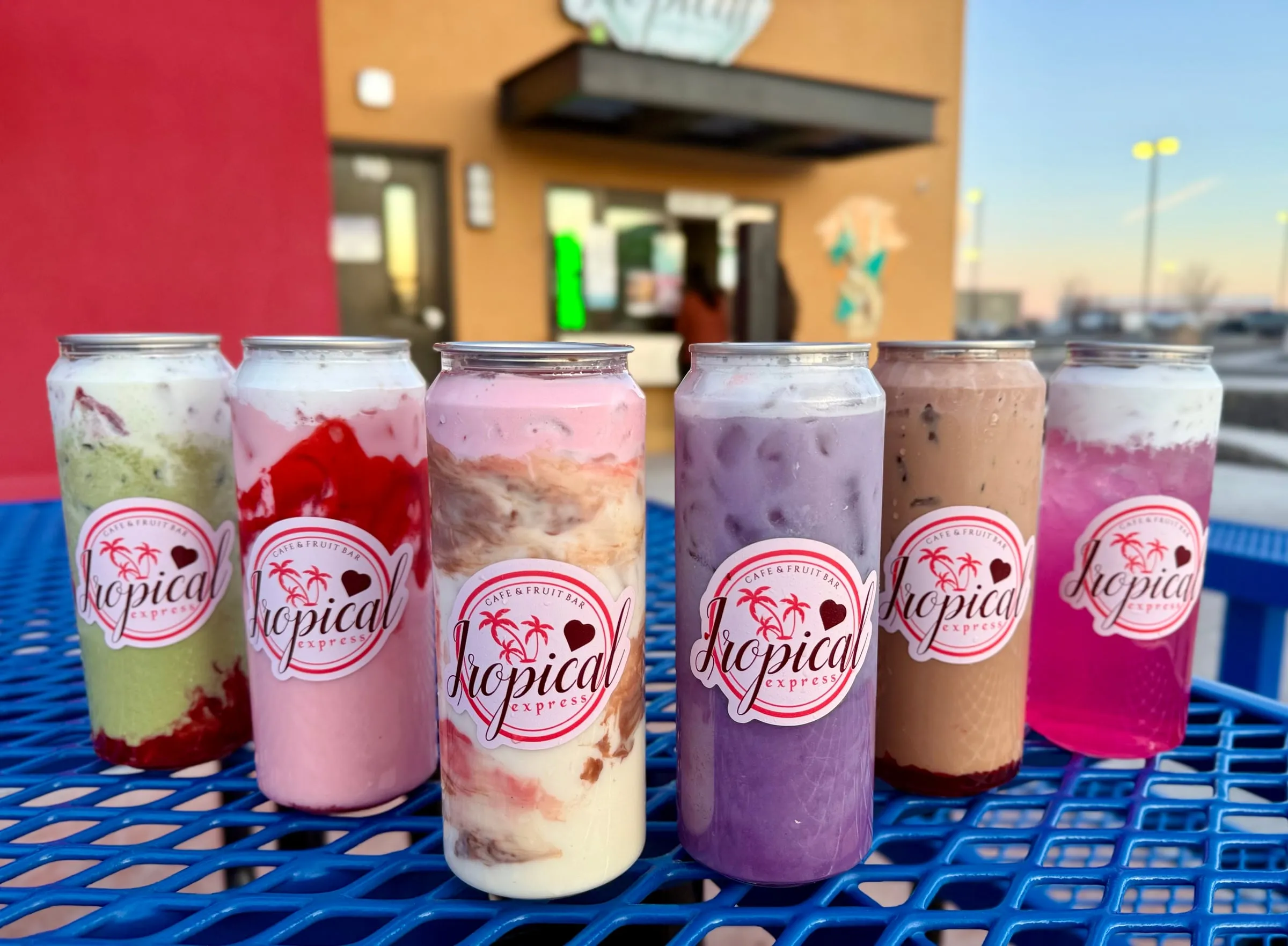 A row of six sealed clear Tropical Express cans on a blue picnic table outside the shop — strawberry matcha, a pink-and-cream refresher, a layered pink drink, a purple drink, mocha, and a magenta refresher — each printed with the palm-tree logo