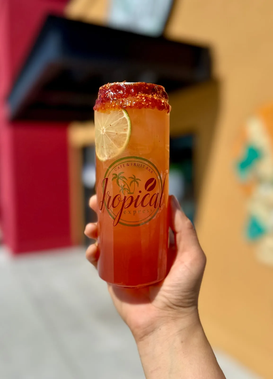 A sealed clear Tropical Express can of a Cantarito — a grapefruit, watermelon and pineapple refresher with a chili-lime chamoy rim dip and a lime wheel — held in front of the storefront