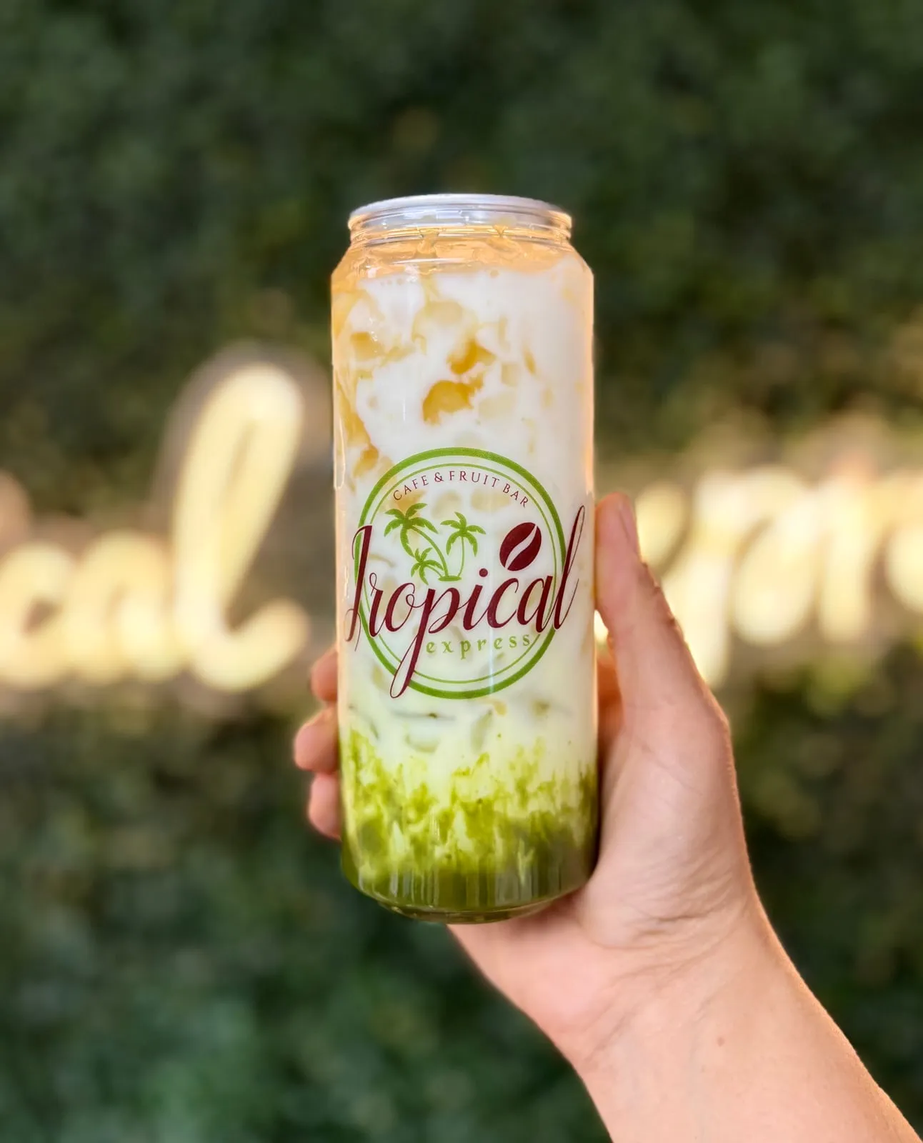 A sealed clear Tropical Express can of a Matcha Macchiato — caramel and milk layered over green matcha settled at the bottom — held in front of greenery and the neon sign