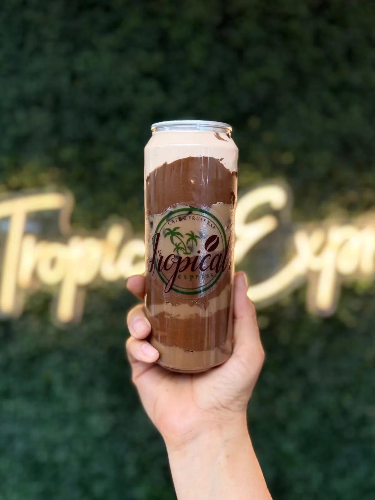 A sealed clear Tropical Express can of a Nutella Latte — a chocolatey Nutella mocha topped with chocolate-hazelnut cold foam — held in front of greenery and the neon sign