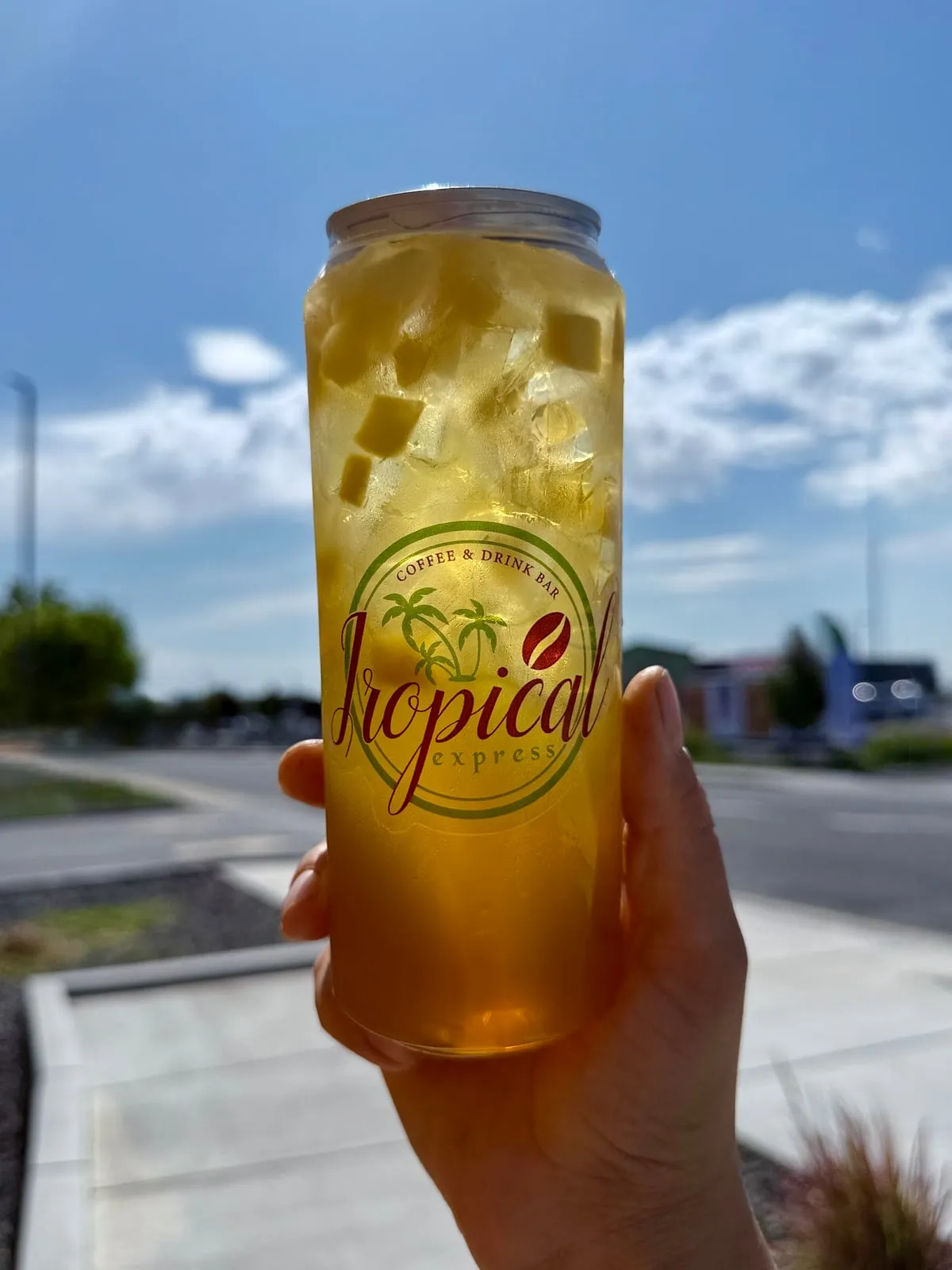 A sealed clear Tropical Express can of a Mango Refresher — golden mango green tea packed with real mango cubes and ice — held up against a bright blue sky