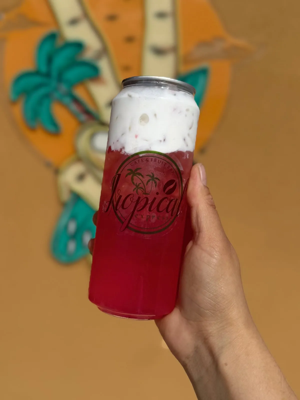 A hand holding a sealed clear Tropical Express can of Pink Paradise — a pink lotus, peach, coconut and guava refresher topped with a thick layer of creamy vanilla cold foam — the palm-tree logo printed on the can, in front of the shop’s painted palm-tree mural