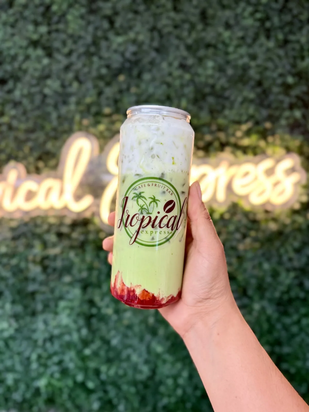 A sealed clear Tropical Express can of Strawberry Matcha — iced green matcha and milk layered over a red strawberry base — held in front of greenery and the glowing neon sign