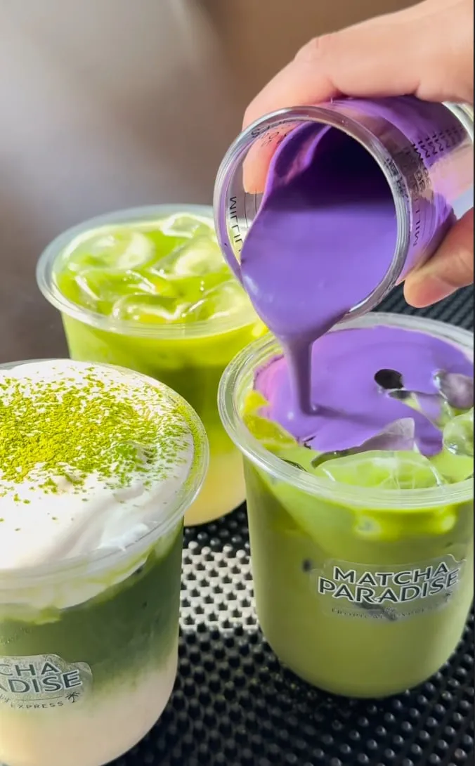 Purple ube cream being poured from a metal pitcher over a cup of iced green matcha at the Matcha Paradise counter, beside a cup topped with matcha-dusted cream