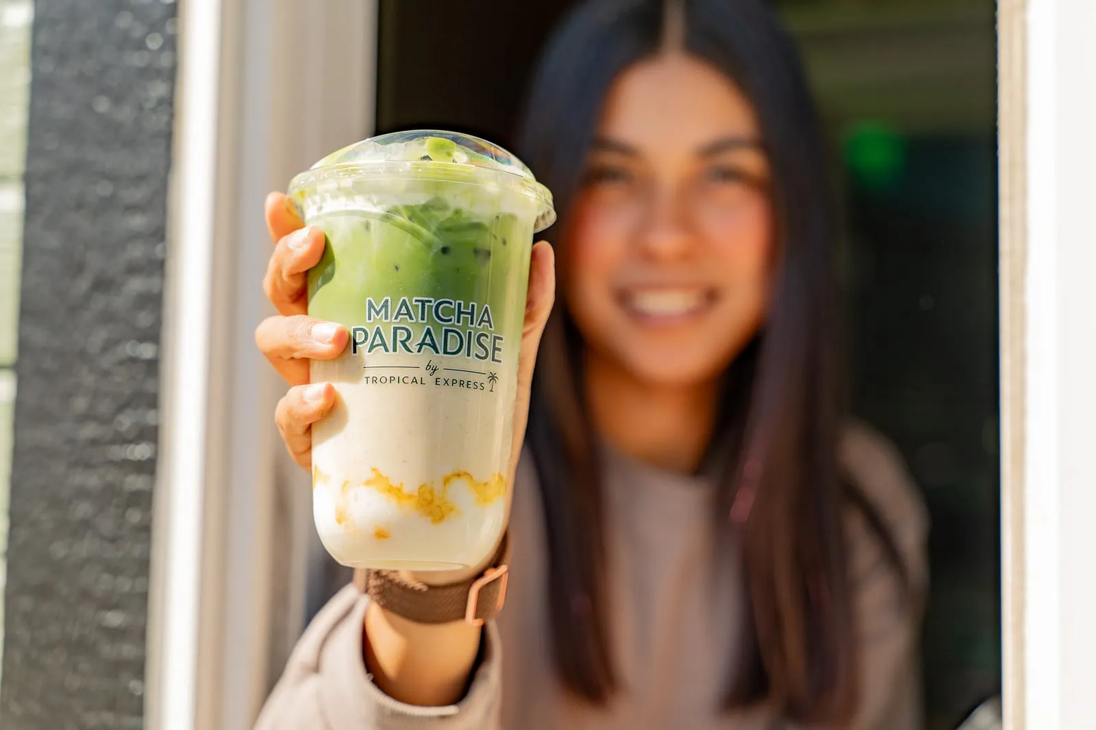 A Mango Sticky Rice Matcha held at the Matcha Paradise service window: golden mango at the bottom, cream in the middle, deep green matcha on top, in a cup printed with the Matcha Paradise logo